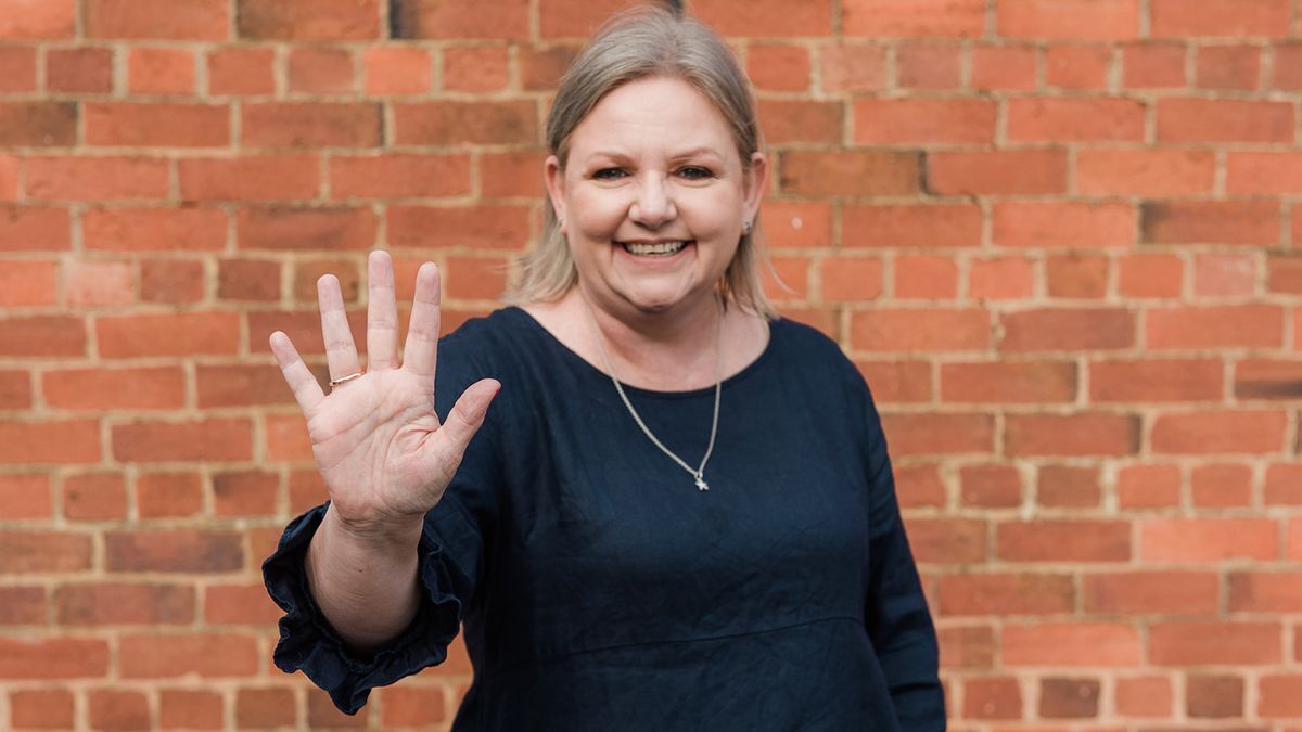 Mell Millgate from Starfish Marketing smiling and holding up five fingers against a red brick wall, representing the five reasons to use video in your marketing.