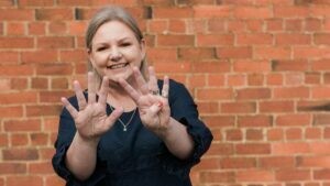 Mell, Founder of Starfish Marketing, smiling, standing in front of a red brick wall, holding up nine fingers to represent nine free marketing tools for small business.