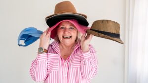 Smiling woman in a pink and white striped shirt playfully wearing several stacked hats, with a blue cap and straw hat on either side, illustrating the challenge of choosing the right marketing platforms.