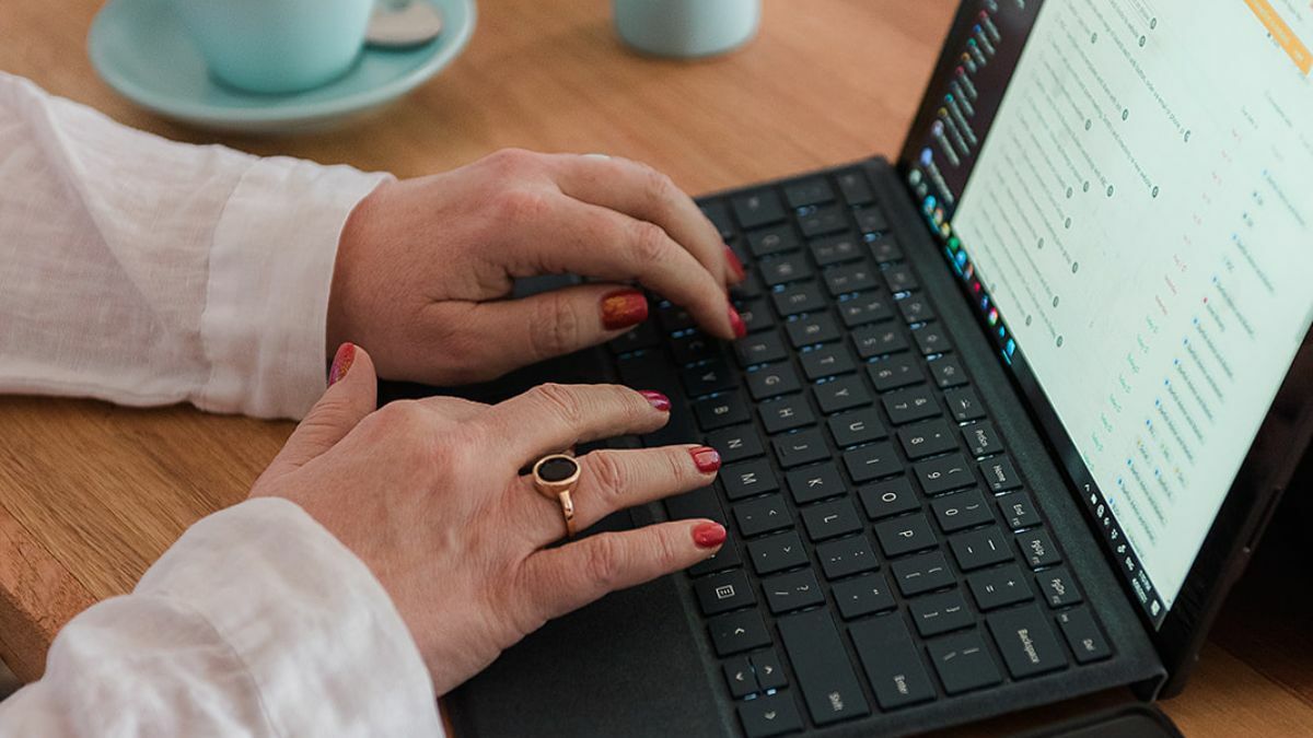 Close-up of a woman’s hands typing on a black laptop keyboard at a wooden desk.