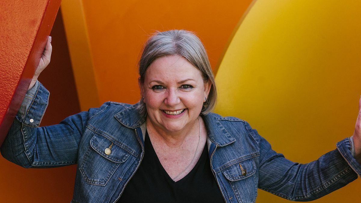 Smiling woman with shoulder-length grey hair wearing a denim jacket stands between two large colourful panels, one red and one yellow. She is looking directly at the camera with a friendly, confident expression. The background is bright and playful, creating a welcoming and energetic vibe.