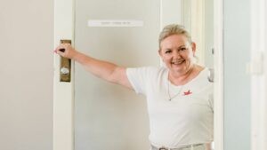 Smiling woman with blonde hair wearing a white t-shirt stands in an open doorway, holding the door with one hand. A small sign on the door reads "open – come on in." She appears warm and welcoming, creating a friendly first impression.