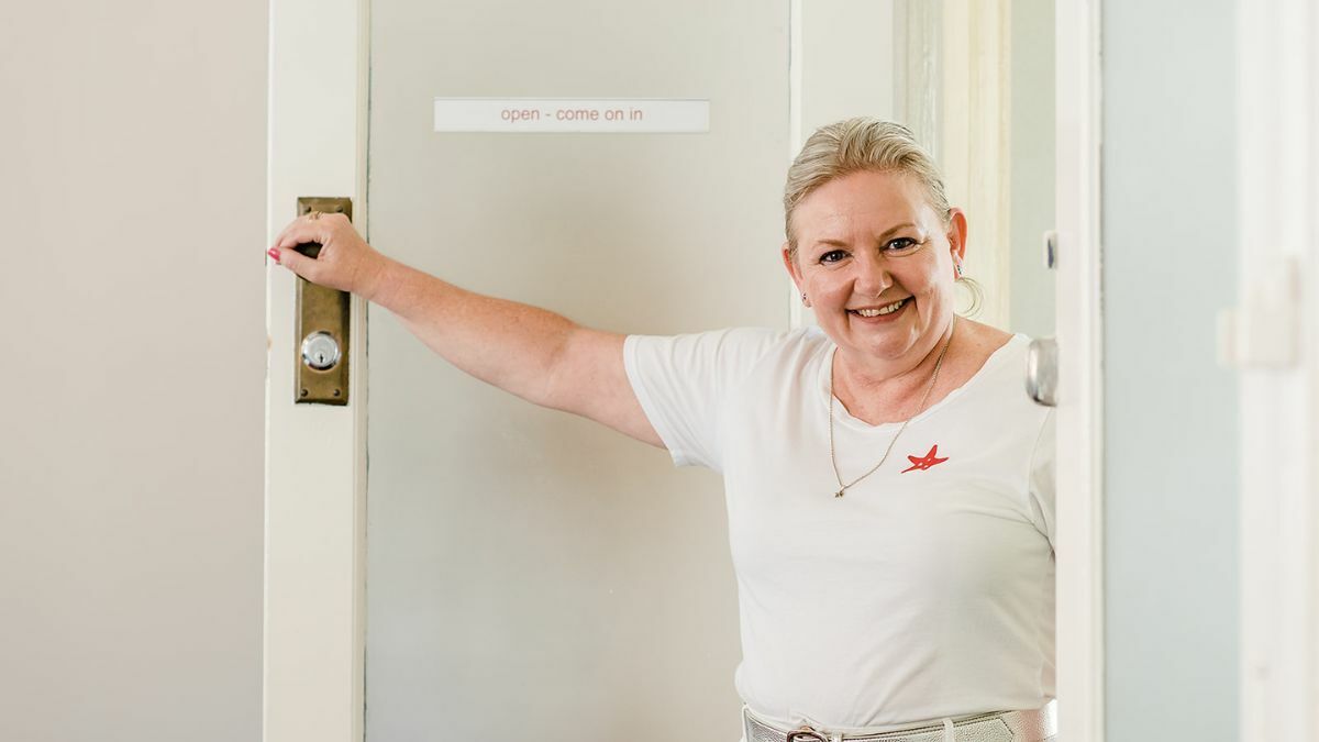 Smiling woman with blonde hair wearing a white t-shirt stands in an open doorway, holding the door with one hand. A small sign on the door reads "open – come on in." She appears warm and welcoming, creating a friendly first impression.