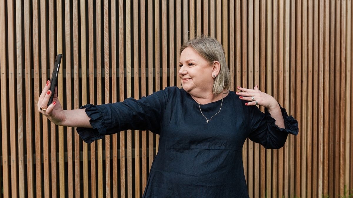 Mell Millgate, a blonde woman and founder of Starfish Marketing in Albury Wodonga, smiling while taking a playful selfie in front of a timber panel wall.