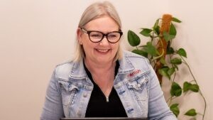 Mell from Starfish Marketing smiling while working on her laptop, wearing a denim jacket with the Starfish Marketing logo, sitting in a bright office with green indoor plants in the background.