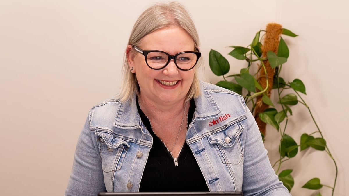 Mell from Starfish Marketing smiling while working on her laptop, wearing a denim jacket with the Starfish Marketing logo, sitting in a bright office with green indoor plants in the background.