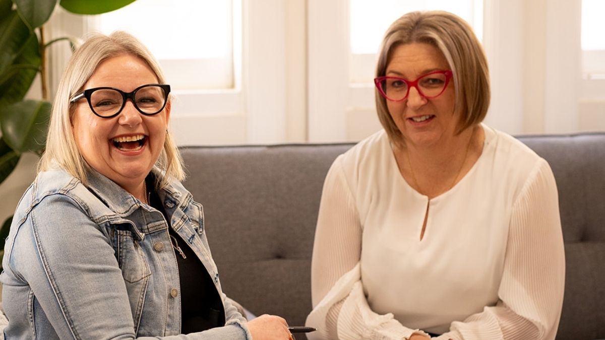 Mell from Starfish Marketing sitting on a grey couch with a client, both smiling and talking during a relaxed marketing strategy meeting in a bright office.