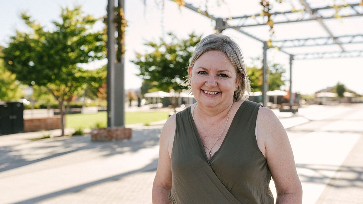 A woman with short blonde hair smiling outdoors in a sunlit public space, standing under a metal pergola structure with trees and paved walkways behind her.