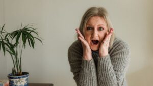 A blonde woman standing indoors with a surprised expression, holding her face with both hands and wearing a grey knitted jumper, with a potted plant beside her.