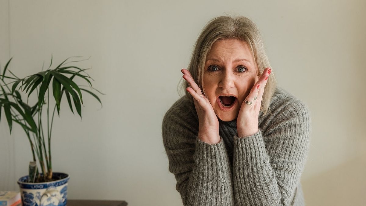 A blonde woman standing indoors with a surprised expression, holding her face with both hands and wearing a grey knitted jumper, with a potted plant beside her.