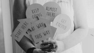 Black and white image of a person holding directional signs reading Start Here, This Way, Keep Going, Almost There and Get Excited, symbolising guidance and consistency in content marketing to build trust.