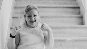 Black and white image of a woman sitting on stairs holding a sign that reads Start Here, smiling and pointing to herself, representing taking the first step in creating content for different platforms.