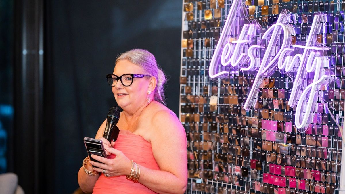 Woman speaking at a business event holding a microphone and phone, with a “Let’s Party” neon sign in the background