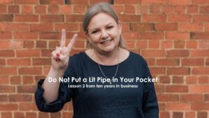 Mell Millgate standing in front of a brick wall holding up two fingers, representing lesson two from ten years in business about thinking before reacting. The words "Do not put a lit pipe in your pocket. Lesson 2 from ten years in business" are overlaid.