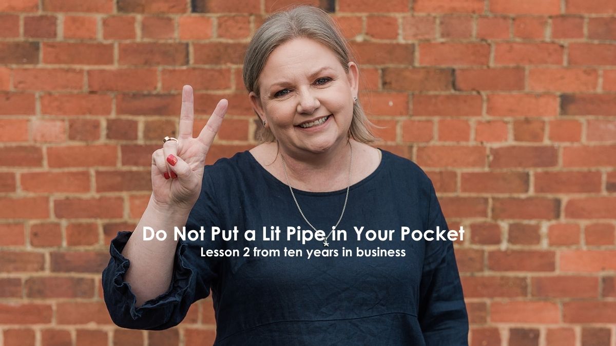 Mell Millgate standing in front of a brick wall holding up two fingers, representing lesson two from ten years in business about thinking before reacting. The words "Do not put a lit pipe in your pocket. Lesson 2 from ten years in business" are overlaid.