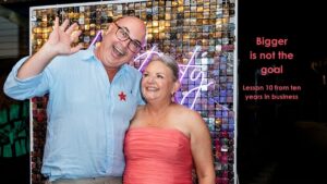 Mell Millgate, founder of Starfish Marketing, smiling with her partner Josh at Starfish Marketing’s ten year celebration. Josh waves beside Mell in front of a reflective Starfish backdrop, with the words “Bigger is not the goal”.