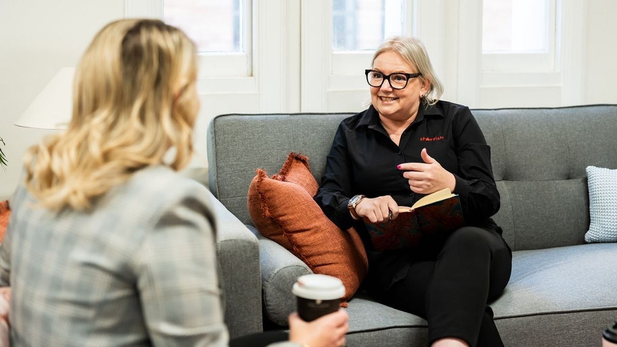 Mell Millgate from Starfish Marketing sitting on a grey sofa, smiling and talking with another woman during a one on one business conversation.